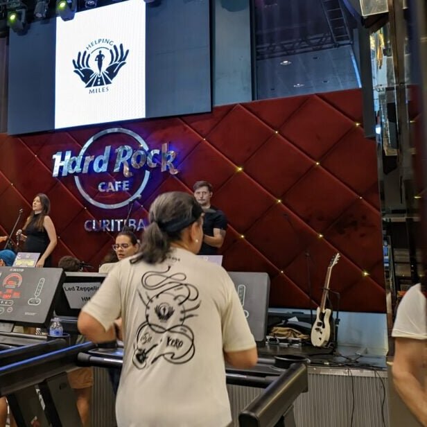 Corredora numa esteira montada no salão do Hard Rock Cafe Curitiba, diante do palco com o letreiro da casa aceso e o telão da Helping Miles