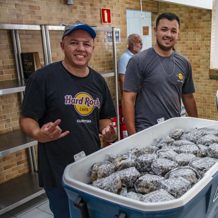 Dois homens da equipe do Hard Rock Cafe Curitiba, um de camiseta da casa, sorriem ao lado de uma caixa térmica cheia de lanches embrulhados, na cozinha do Mesa Solidária