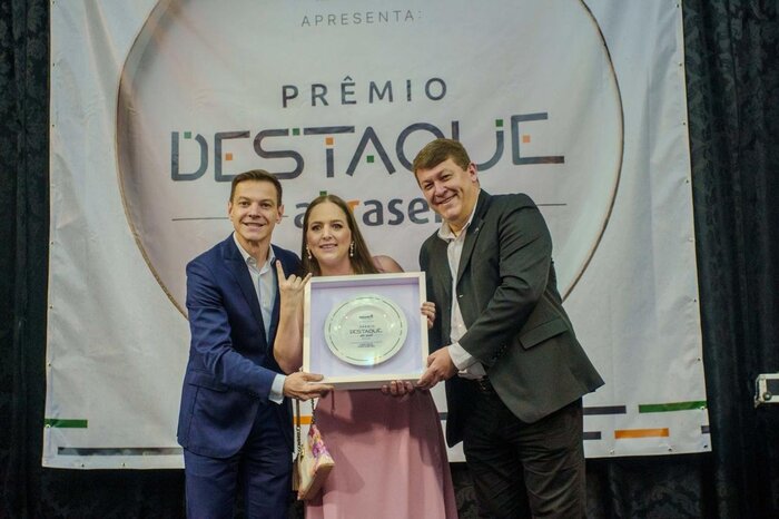 Três pessoas no palco seguram o troféu do Prêmio Destaque Abrasel, um prato emoldurado, diante do painel do prêmio